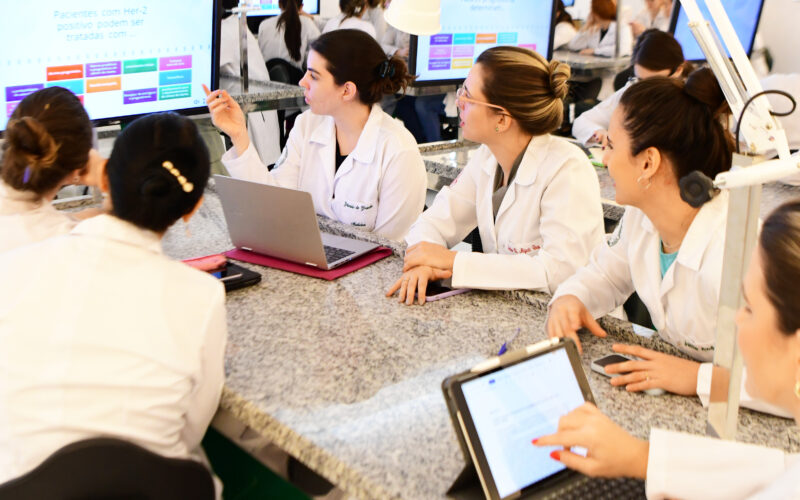 Estudantes de Medicina da Unoeste em um ambiente de aprendizado interativo, usando laptops e tablets enquanto discutem em grupo, com telas ao fundo exibindo conteúdos relacionados ao tratamento de pacientes com Her-2 positivo.