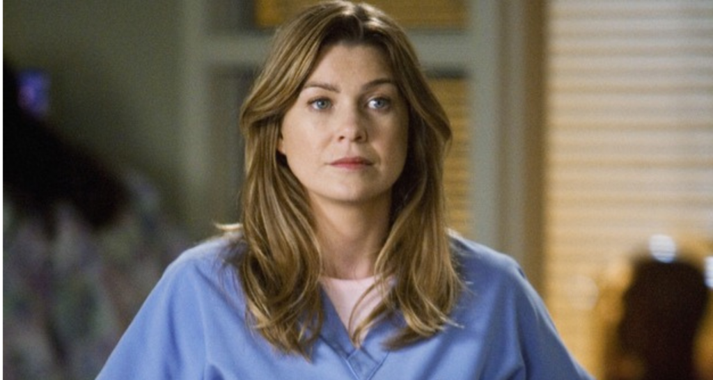 Um close-up de Ellen Pompeo como Meredith Grey em Grey's Anatomy. Ela está usando um uniforme azul e olhando para a câmera com uma expressão séria.