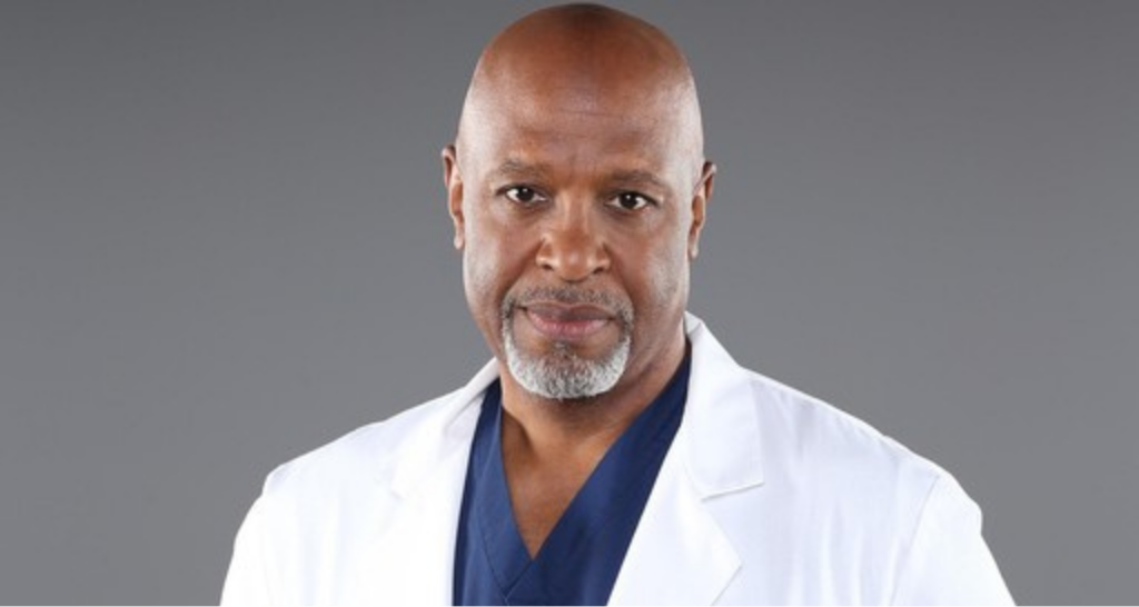 Um retrato de James Pickens Jr. como Richard Webber em Grey's Anatomy. Ele está usando um jaleco branco sobre um uniforme azul marinho e olhando para a câmera com uma expressão neutra. Ele tem a cabeça raspada e uma barba grisalha. O fundo é cinza sólido.