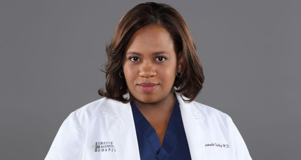Um retrato de Chandra Wilson como Miranda Bailey em Grey's Anatomy. Ela está usando um jaleco branco sobre um uniforme azul marinho e olhando para a câmera com uma expressão neutra. O fundo é cinza sólido.