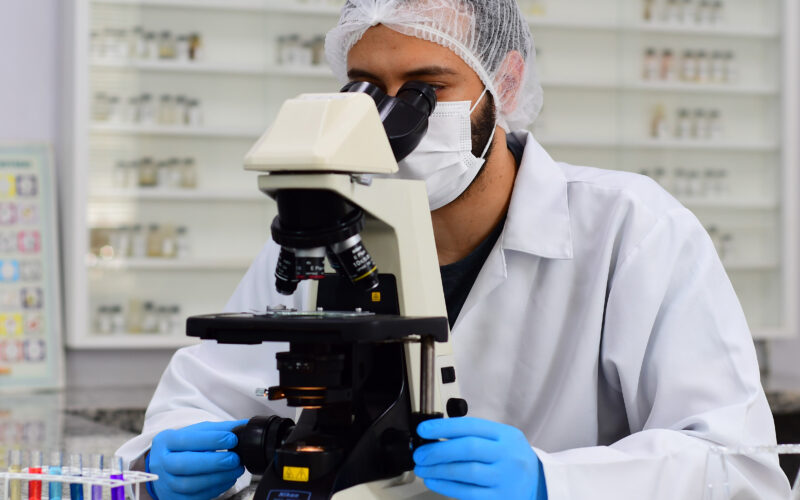 Homem de jaleco branco, luvas azuis, máscara e touca de proteção, observando amostras em um microscópio dentro de um laboratório. Ao fundo, há frascos e materiais de pesquisa organizados em prateleiras.