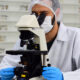 Homem de jaleco branco, luvas azuis, máscara e touca de proteção, observando amostras em um microscópio dentro de um laboratório. Ao fundo, há frascos e materiais de pesquisa organizados em prateleiras.
