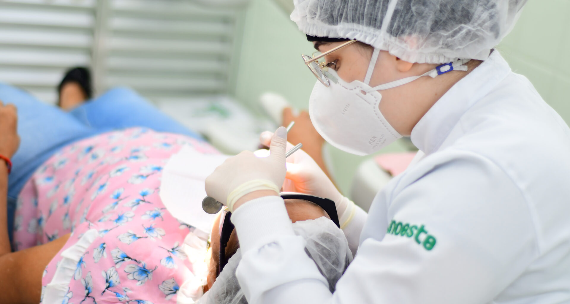 Estudante de Odontologia da Unoeste realizando um atendimento odontológico em um paciente, utilizando equipamentos de proteção individual (EPI), como máscara, touca e luvas, em um ambiente clínico.