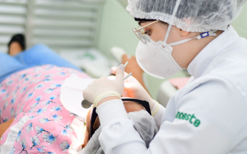 Estudante de Odontologia da Unoeste realizando um atendimento odontológico em um paciente, utilizando equipamentos de proteção individual (EPI), como máscara, touca e luvas, em um ambiente clínico.