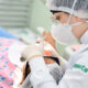 Estudante de Odontologia da Unoeste realizando um atendimento odontológico em um paciente, utilizando equipamentos de proteção individual (EPI), como máscara, touca e luvas, em um ambiente clínico.