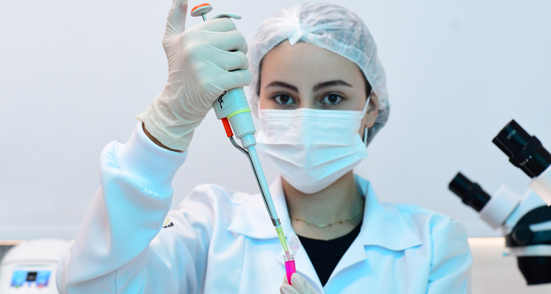 Estudante de Biomedicina vestindo jaleco branco, máscara e touca de proteção, manipulando uma micropipeta em um laboratório, com microscópios ao fundo.