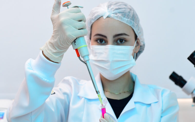 Estudante de Biomedicina vestindo jaleco branco, máscara e touca de proteção, manipulando uma micropipeta em um laboratório, com microscópios ao fundo.