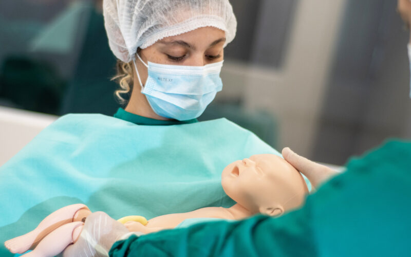 Estudante de Medicina da Unoeste treinando procedimentos de parto em um manequim neonatal, utilizando equipamentos de proteção, como máscara e touca, em um ambiente simulado de saúde.