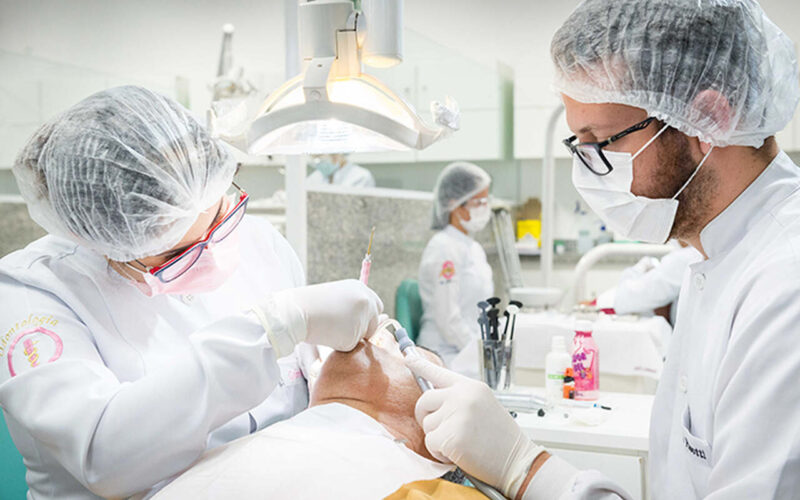 A imagem mostra dois estudantes de Odontologia na clínica-escola Unoeste atendendo um paciente. O paciente está deitado em uma cadeira de dentista e os dentistas estão usando máscaras e luvas. A aluna da esquerda está usando touca, óculos e máscara, e está segurando um instrumento na mão direita. O aluno da direita também está usando máscara, touca e óculos e está segurando um instrumento na mão esquerda.