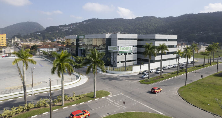 Imagem do campus da Unoeste no Guarujá, São Paulo, com uma grande estrutura moderna de dois prédios espelhados, cercada por palmeiras ao longo das vias. No primeiro plano, uma rua com carros circulando e ao fundo uma paisagem de montanhas verdes e edifícios residenciais.