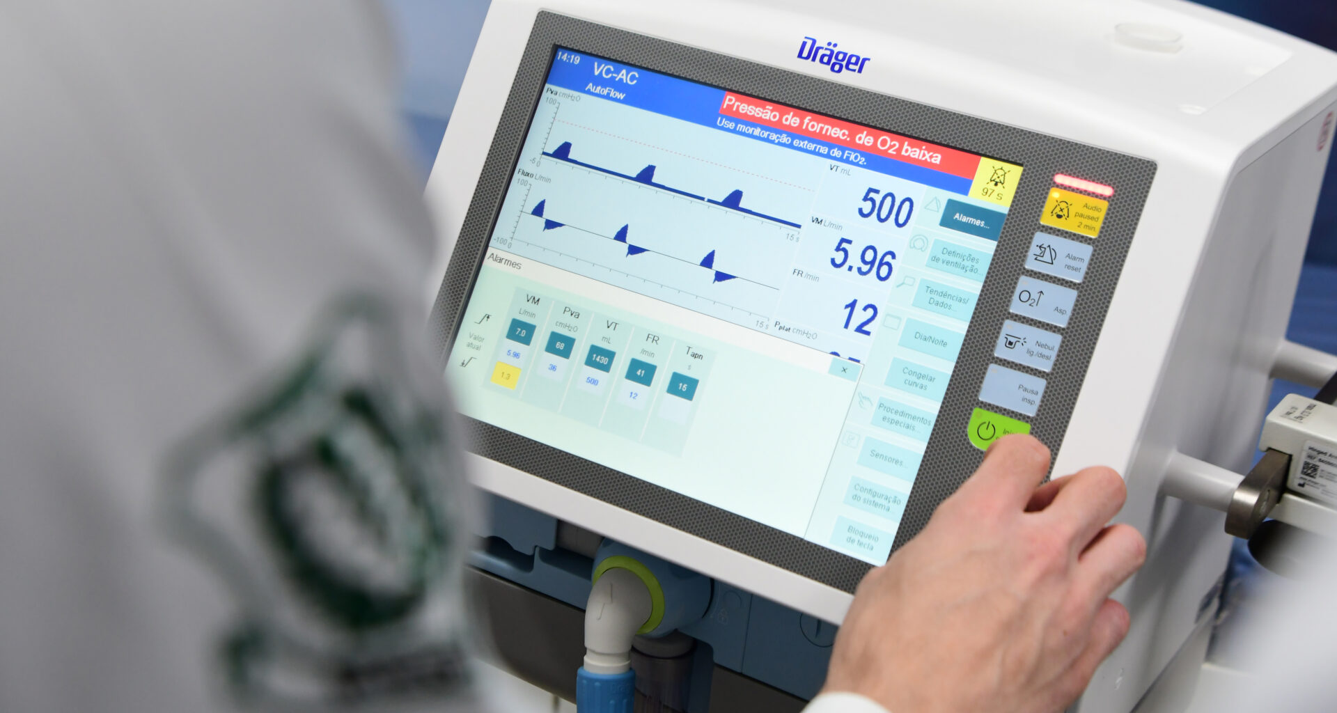 Fotografia de um ventilador mecânico moderno da marca Dräger, exibindo uma tela digital com parâmetros detalhados de ventilação, como pressão, volume e frequência respiratória. A mão de um estudante de medicina da Unoeste aparece na imagem ajustando o dispositivo, que está em uso em um laboratório de aula prática do curso.