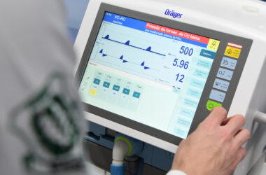 Fotografia de um ventilador mecânico moderno da marca Dräger, exibindo uma tela digital com parâmetros detalhados de ventilação, como pressão, volume e frequência respiratória. A mão de um estudante de medicina da Unoeste aparece na imagem ajustando o dispositivo, que está em uso em um laboratório de aula prática do curso.
