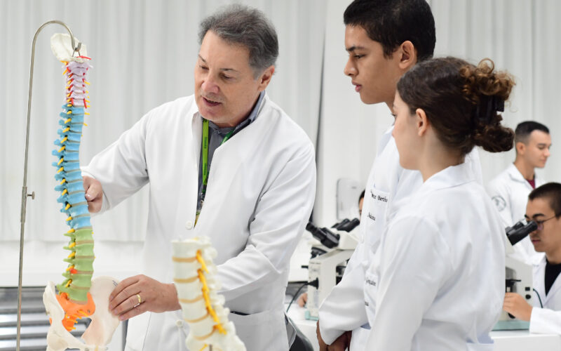 professor da Unoeste de jaleco branco explicando a estrutura da coluna vertebral em um modelo anatômico para três estudantes de medicina, que estão atentos à demonstração em um ambiente de laboratório, com microscópios ao fundo.