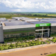 Imagem aérea do campus da Unoeste em Jaú - SP, destacando a fachada moderna do edifício com painéis solares no teto, cercado por áreas verdes e um amplo estacionamento, em um ambiente rural com plantações ao fundo.