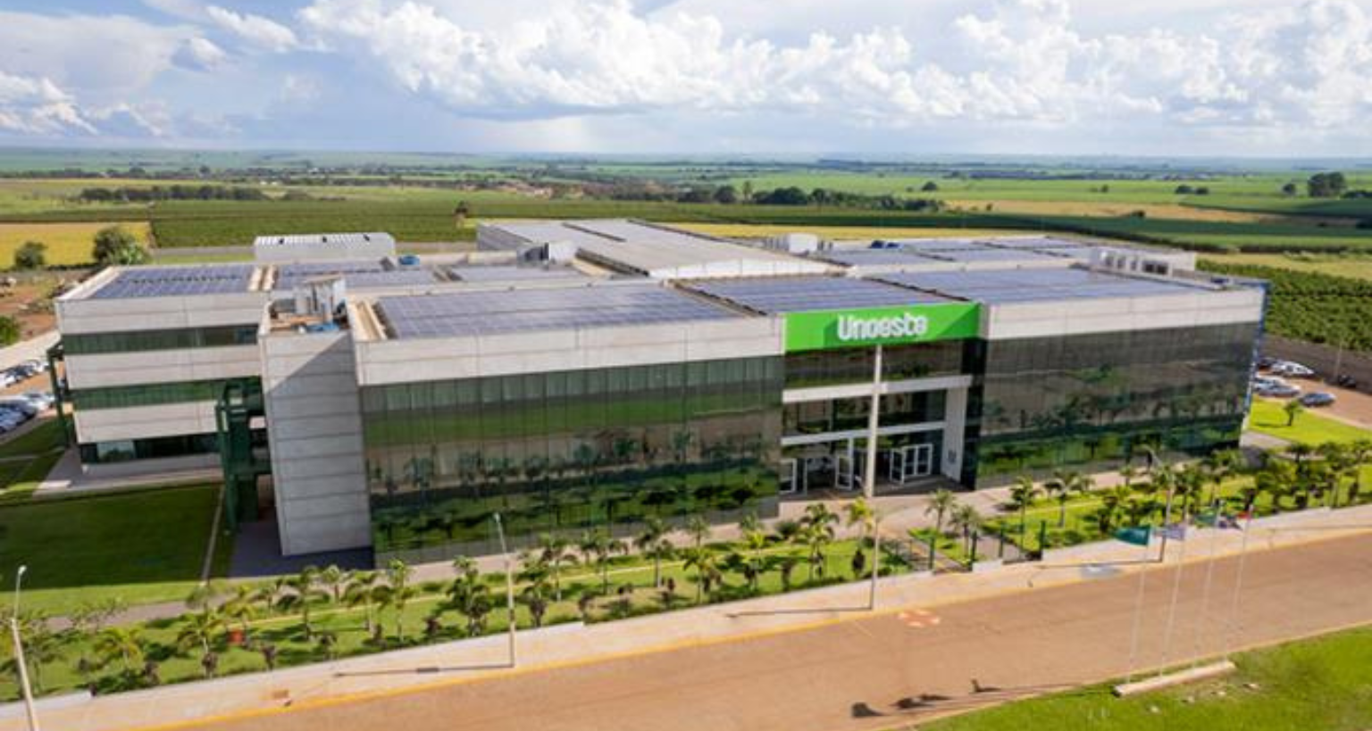 Imagem aérea do campus da Unoeste em Jaú - SP, destacando a fachada moderna do edifício com painéis solares no teto, cercado por áreas verdes e um amplo estacionamento, em um ambiente rural com plantações ao fundo.