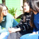 Três jovens mulheres sentadas em um banco em um parque, conversando. A mulher à esquerda usa uma blusa verde-água de mangas compridas e calças brancas. Ela usa óculos e tem cabelos castanhos claros e compridos. A mulher no centro usa uma blusa listrada em preto e branco e calças escuras. Ela tem cabelos castanhos escuros e compridos. A mulher à direita usa uma blusa azul de mangas compridas e uma mochila bege com detalhes em marrom. Ela tem cabelos castanhos escuros e compridos. Ao fundo, vê-se grama verde, uma pessoa em uma motocicleta e uma parede verde. A cena sugere um dia ensolarado em uma cidade do interior de São Paulo.