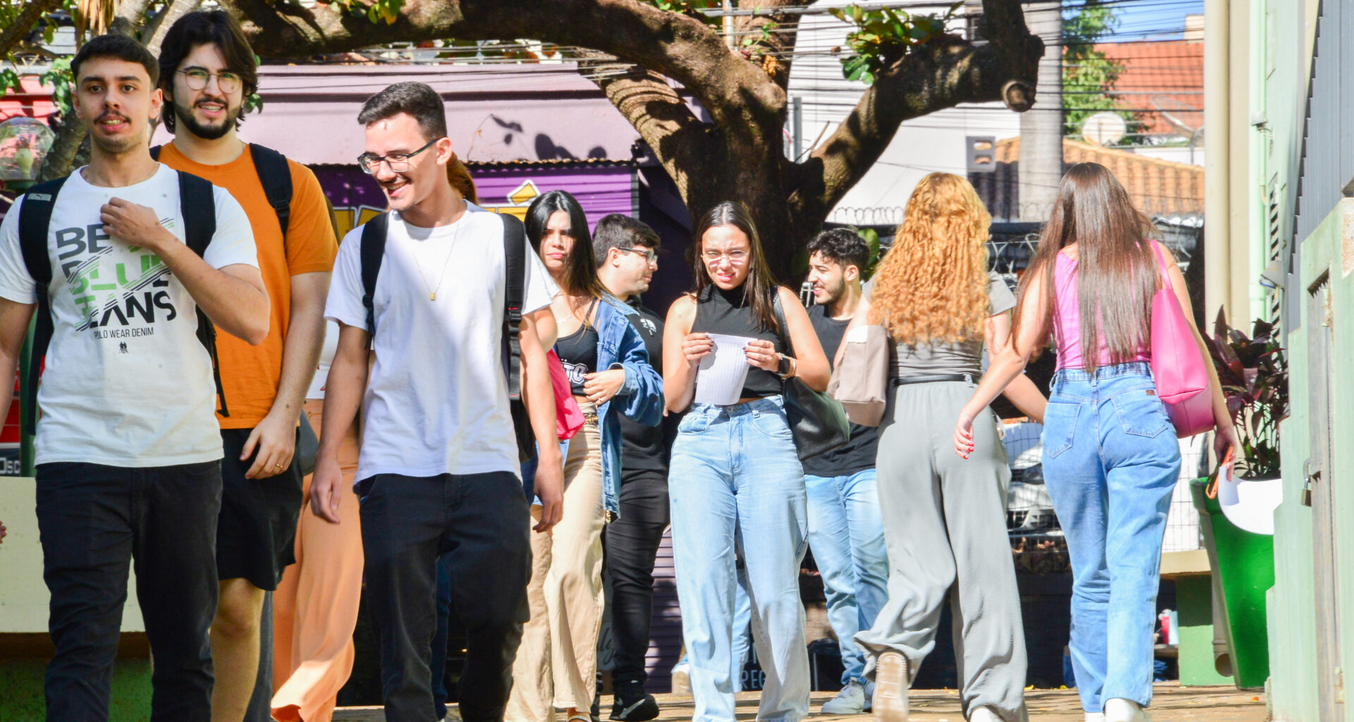 Grupo de estudantes caminha alegremente em uma área arborizada de um campus universitário. Eles carregam mochilas e interagem entre si em um ambiente descontraído. Ao fundo, é possível observar árvores, prédios e detalhes que reforçam o clima acolhedor e vibrante do espaço acadêmico.
