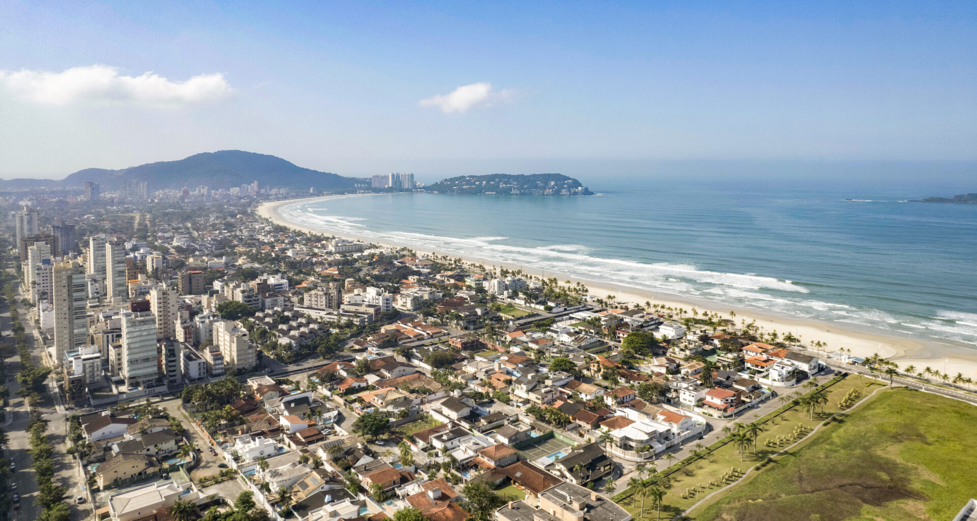 Vista aérea de Guarujá, destacando a Praia da Enseada com sua extensa faixa de areia e mar azul. A imagem revela a combinação de áreas residenciais e prédios à beira-mar, evidenciando o estilo de vida praiano da região.
