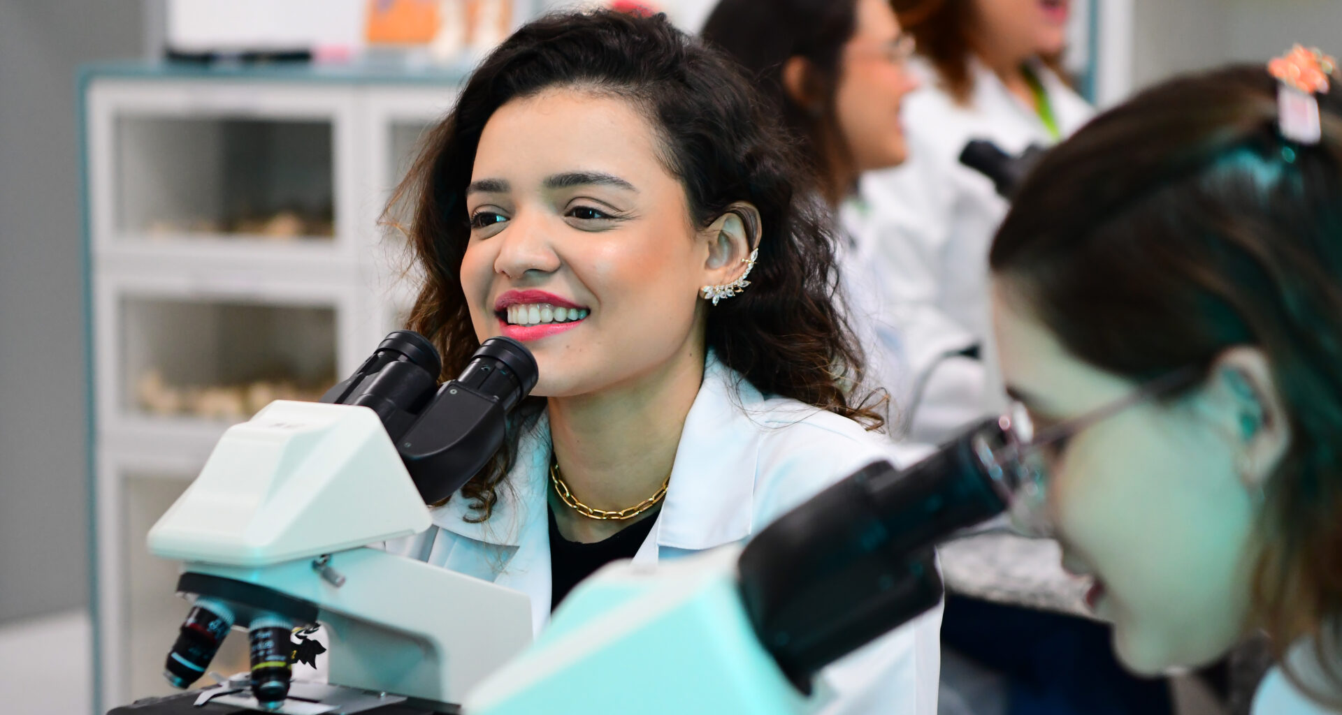 Estudantes de biomedicina da Unoeste em um laboratório, vestindo jalecos brancos e utilizando microscópios. Em destaque, uma jovem sorridente observa pelo equipamento, demonstrando entusiasmo pela prática científica.