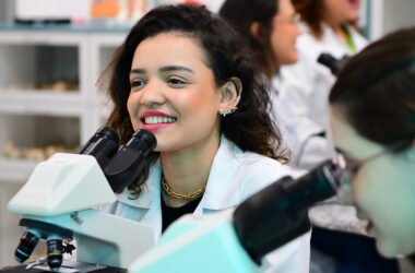 Estudantes de biomedicina da Unoeste em um laboratório, vestindo jalecos brancos e utilizando microscópios. Em destaque, uma jovem sorridente observa pelo equipamento, demonstrando entusiasmo pela prática científica.