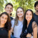 Grupo de cinco jovens estudantes sorridentes em um ambiente externo, com árvores e outras pessoas ao fundo. Eles estão vestidos de forma casual e parecem felizes, representando diversidade e interação em um ambiente estudantil.