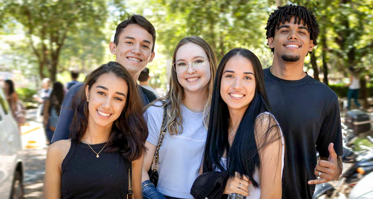 Grupo de cinco jovens estudantes sorridentes em um ambiente externo, com árvores e outras pessoas ao fundo. Eles estão vestidos de forma casual e parecem felizes, representando diversidade e interação em um ambiente estudantil.