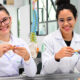 Duas estudantes de medicina da Unoeste, vestindo jalecos brancos, sorriem enquanto seguram pequenas peças anatômicas em uma sala de aula prática. Ao fundo, um modelo de coluna vertebral e crânios auxiliam no estudo da anatomia. A sala é bem iluminada, e elas parecem engajadas no aprendizado.