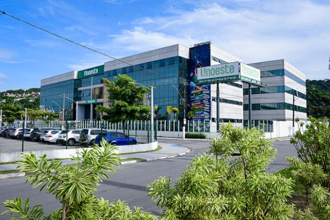 O prédio da Unoeste (Universidade do Oeste Paulista), campus Guarujá, visto em um dia ensolarado com céu azul. O edifício moderno de vários andares, com fachada de vidro e painéis brancos, exibe placas com a marca "Unoeste". Um estacionamento com vários carros é visível em frente ao prédio. Arbustos verdes e folhagens baixas adornam o primeiro plano, adicionando um toque de natureza à cena urbana. Uma rua curva passa em frente ao prédio, levando o olhar do espectador em direção à estrutura.

