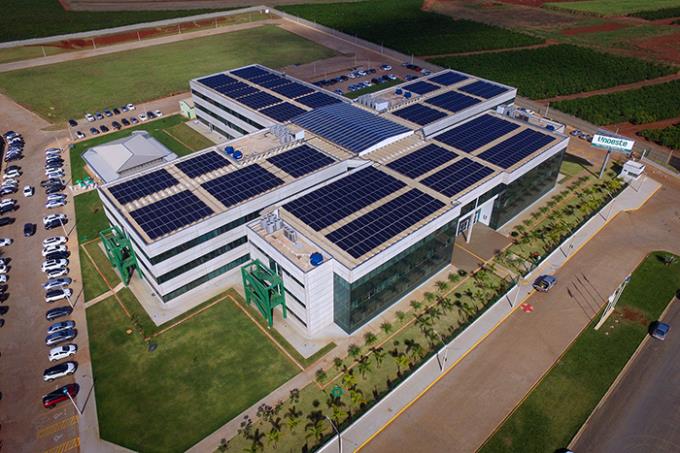 Uma vista aérea do campus da Unoeste (Universidade do Oeste Paulista) em Jaú, São Paulo. A imagem destaca o telhado do prédio principal coberto por painéis solares, demonstrando o compromisso da universidade com a sustentabilidade. Vemos também um amplo estacionamento com diversos carros e áreas verdes ao redor do edifício, além de uma rua que passa ao lado do campus. Ao fundo, é possível observar uma área rural, provavelmente campos de cultivo. A imagem, feita em um dia claro e ensolarado, oferece uma perspectiva abrangente da infraestrutura e localização do campus.

