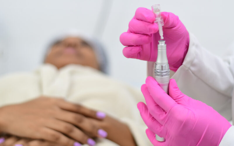 Profissional da área da saúde, vestindo luvas rosas e jaleco branco, segura um dispositivo utilizado em procedimentos estéticos. Ao fundo, uma paciente deitada na maca aguarda o início do tratamento.