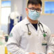 A imagem mostra um estudante de Medicina em um ambiente hospitalar, vestindo um jaleco branco com o emblema da Unoeste no braço e um crachá de identificação no bolso. Ele usa óculos, máscara cirúrgica e tem um estetoscópio pendurado no pescoço. O estudante segura um papel e está posicionado em um cenário clínico, com equipamentos médicos e leitos ao fundo, transmitindo a atmosfera de aprendizado prático do internato médico.