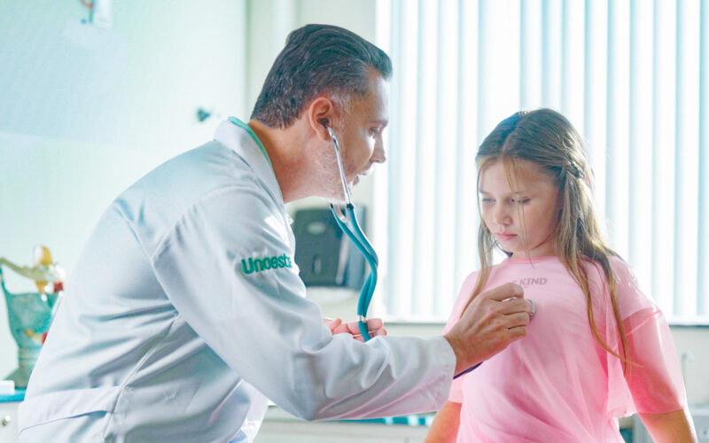 Médico com jaleco da Unoeste utilizando um estetoscópio para examinar uma menina com blusa rosa em um consultório iluminado, com modelos anatômicos ao fundo.
