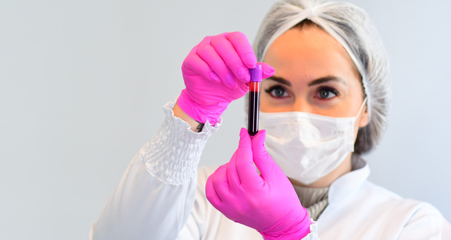 Uma estudante de biomedicina da Unoeste, vestindo jaleco branco, touca, máscara facial e luvas, segurando um frasco com material de coleta laboratorial.