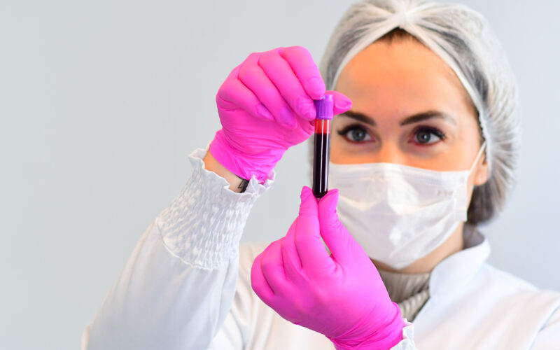 Uma estudante de biomedicina da Unoeste, vestindo jaleco branco, touca, máscara facial e luvas, segurando um frasco com material de coleta laboratorial.