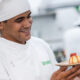 Um estudante de gastronomia, vestido com uniforme branco e toque blanche, segura um prato com uma sobremesa delicadamente decorada. Ele sorri enquanto admira sua criação, demonstrando orgulho e dedicação. Ao fundo, outros alunos trabalham em uma cozinha profissional, reforçando o ambiente de aprendizado prático. O uniforme exibe o nome "Unoeste", indicando a instituição de ensino.