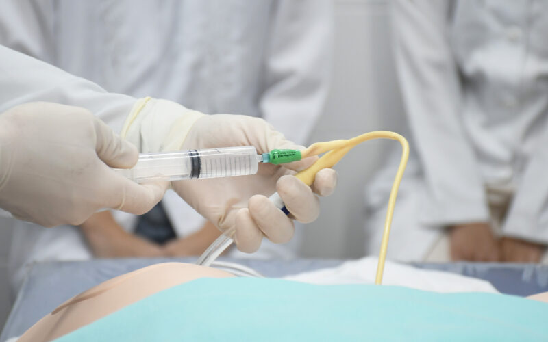 Mãos enluvadas de um profissional de saúde manipulam uma sonda e uma seringa durante um procedimento médico simulado em um ambiente hospitalar. Ao fundo, estudantes ou profissionais da área observam atentamente a demonstração.
