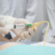 Mãos enluvadas de um profissional de saúde manipulam uma sonda e uma seringa durante um procedimento médico simulado em um ambiente hospitalar. Ao fundo, estudantes ou profissionais da área observam atentamente a demonstração.