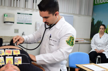 Estudante de Medicina realiza exame físico com estetoscópio em paciente durante aula prática, com supervisão docente em laboratório de simulação clínica.