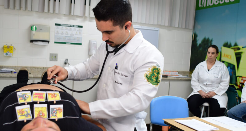 Estudante de Medicina realiza exame físico com estetoscópio em paciente durante aula prática, com supervisão docente em laboratório de simulação clínica.