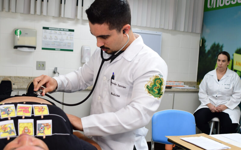 Estudante de Medicina realiza exame físico com estetoscópio em paciente durante aula prática, com supervisão docente em laboratório de simulação clínica.