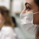 Rosto de uma profissional de saúde com olhar atento utilizando máscara facial descartável em um ambiente hospitalar. Ao fundo, estudantes ou profissionais da área também dividem o local.