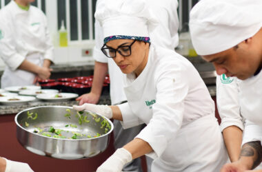 Estudantes do curso de Gastronomia da Unoeste preparam e finalizam pratos em aula prática de cozinha, vestindo uniformes completos e atuando em equipe.