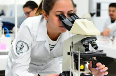 Aluna do curso de Medicina da Unoeste observa amostra em microscópio durante aula prática em laboratório moderno, usando jaleco com o brasão da faculdade de Medicina de Presidente Prudente.
