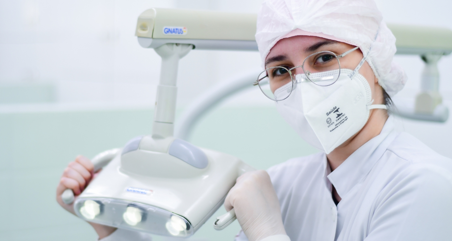 Estudante de Odontologia com jaleco e máscara segura refletor odontológico em clínica-escola, representando a prática profissional durante a faculdade.