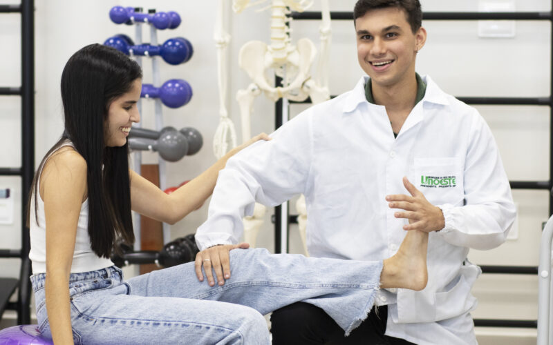 Estudante do curso de Fisioterapia da Unoeste realiza atendimento simulado com paciente em ambiente de laboratório, utilizando bola de exercícios e recursos de apoio ao fundo.