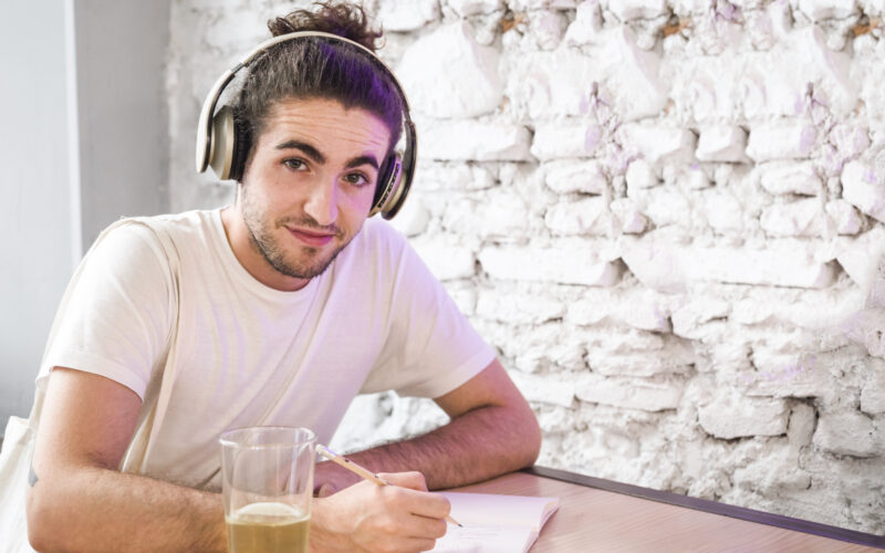 Jovem com coque estudando com fones de ouvido enquanto escuta podcasts para aprender inglês. Copo com líquido amarelado ao lado, fundo da parede branca texturizada.