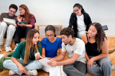 A imagem mostra um grupo de sete jovens adultos (quatro mulheres e três homens) com aparências diversas, sentados de forma descontraída em degraus largos de madeira compensada clara (estilo arquibancada). O ambiente é minimalista, com uma parede cinza-clara ao fundo, criando uma atmosfera de sala de aula moderna, espaço de coworking ou área de convivência universitária.