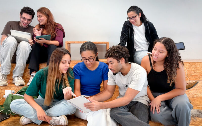 A imagem mostra um grupo de sete jovens adultos (quatro mulheres e três homens) com aparências diversas, sentados de forma descontraída em degraus largos de madeira compensada clara (estilo arquibancada). O ambiente é minimalista, com uma parede cinza-clara ao fundo, criando uma atmosfera de sala de aula moderna, espaço de coworking ou área de convivência universitária.