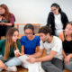 A imagem mostra um grupo de sete jovens adultos (quatro mulheres e três homens) com aparências diversas, sentados de forma descontraída em degraus largos de madeira compensada clara (estilo arquibancada). O ambiente é minimalista, com uma parede cinza-clara ao fundo, criando uma atmosfera de sala de aula moderna, espaço de coworking ou área de convivência universitária.