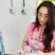 Médica residente com jaleco rosa registra informações em prontuário ao lado de equipamento hospitalar, representando a rotina de plantão na área da saúde.